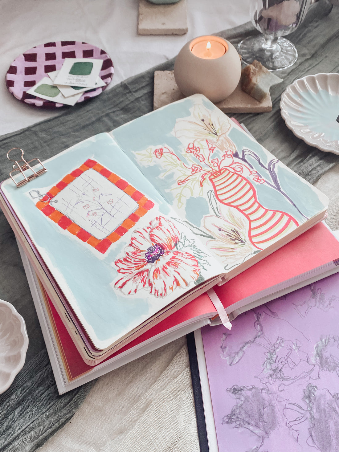 Open artist's sketchbook on a table showing a drawing of a vase with flowers, set against a blue background. 