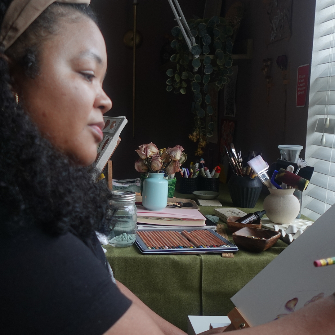 artist holding a paintbrush at her easel with an art table in the background, displaying flowers, paintbrushes, and art supplies.